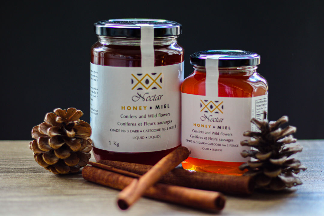 Nectar honey jars of 1Kg and 500g seating the one beside the other on a countertop. Cinammon and pine cones decoration. Black background.